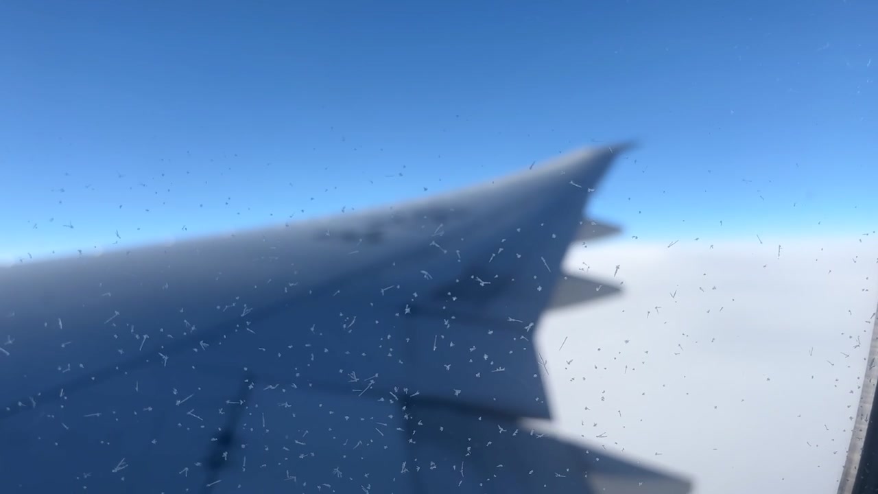 View from an airplane window showing the wing above a layer of white clouds. Clear blue sky and water droplets on the window appear during flight over Russia.