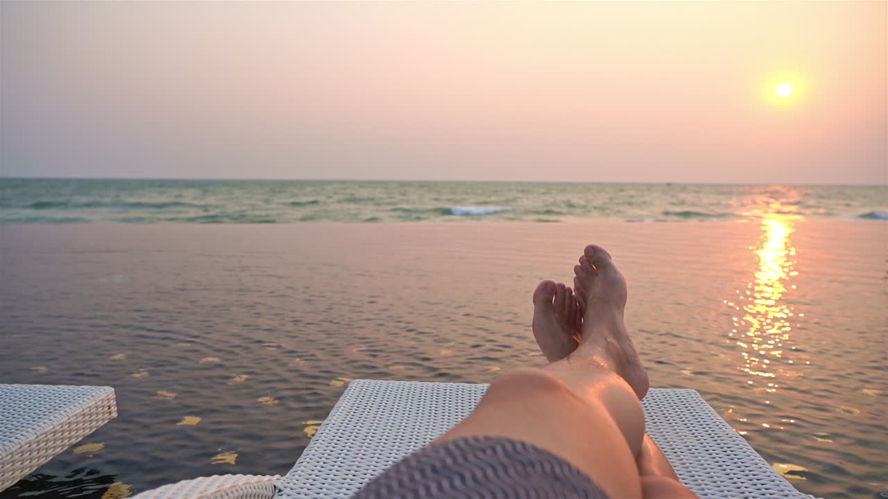 Vista en primera persona desde una tumbona al borde de la piscina de un resort, disfrutando de la vista del atardecer sobre el océano.