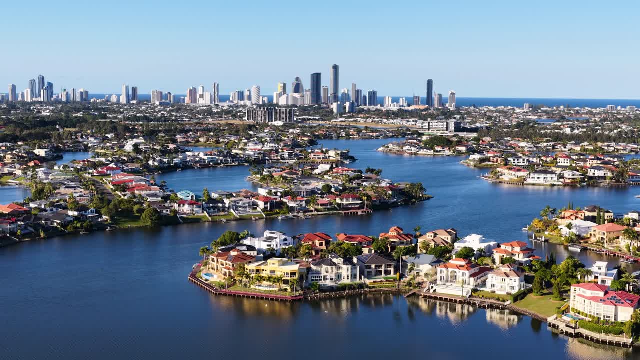 Drone camera glides above riverfront residential estates toward Gold Coast skyline under clear daylight, revealing urban landscape, waterways, and luxury homes