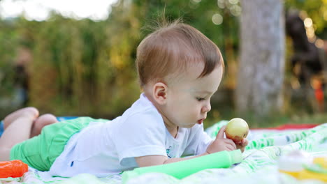 having rest in the summer garden. adorable baby boy lies on the belly looking attentively at the apple. blurred backdrop.