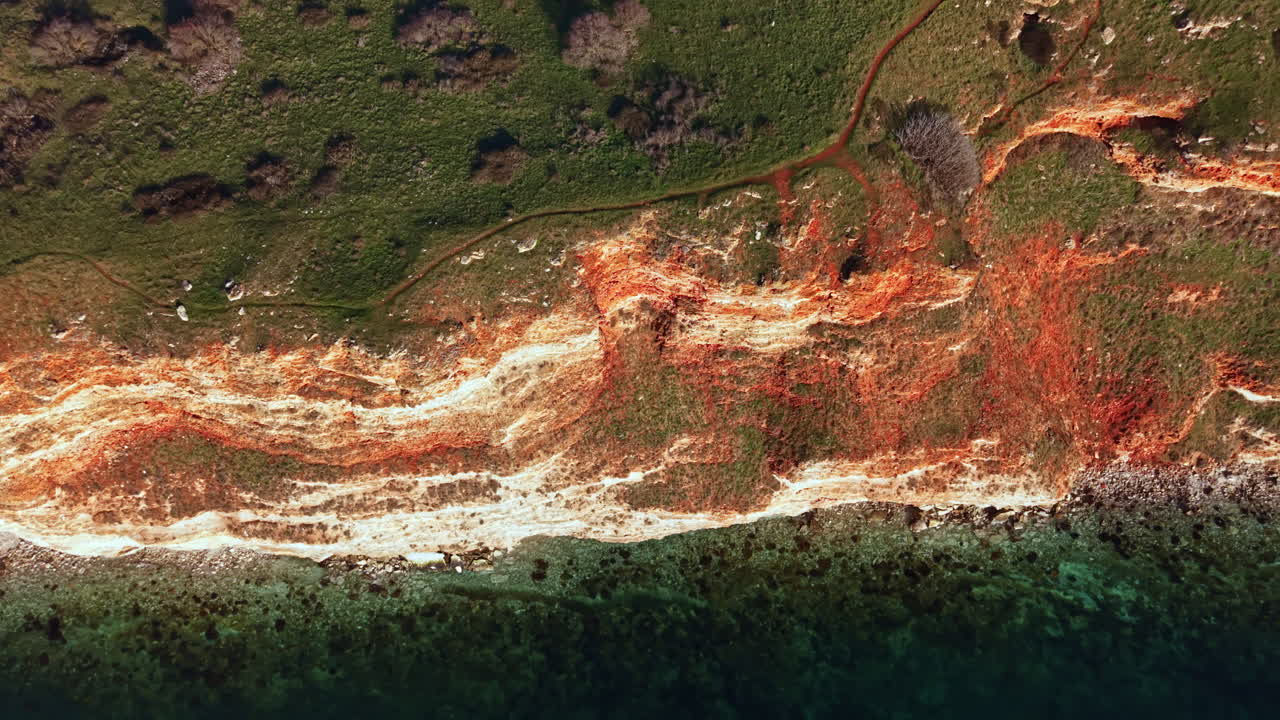 Coastline view showing layers of earth by water during daytime with green area above and rocky shore below near sea