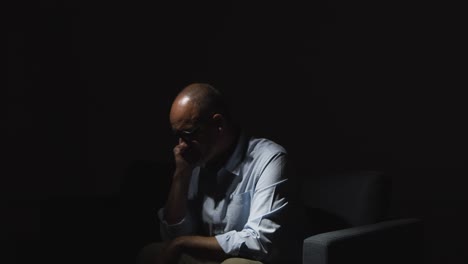 우울증에 걸린 성숙한 남자가 정신 건강에 어려움을 겪고 어 ⁇  속에서 소파에 앉아 낮은 조명을 받고 있습니다.