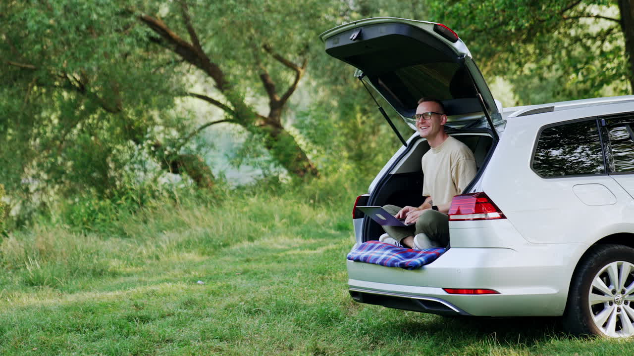 Happy man sits in his car trunk in the forest. Smiling freelancer works on laptop enjoying nature.