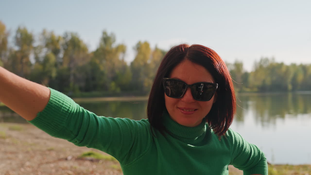 Donna che tiene un'amaca arrotolata in riva al lago, che invita al riposo e all'allestimento, sorridente con occhiali da sole e un maglione verde; acqua calma e linea di alberi sullo sfondo, atmosfera soleggiata in riva al lago e gesto casuale da campeggio.