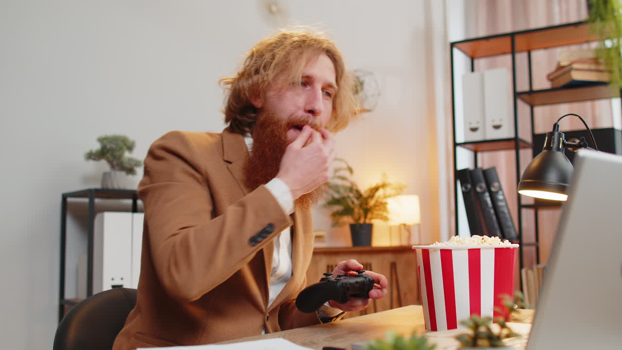 Businessman eating popcorn while playing video game on laptop spending leisure time at home office