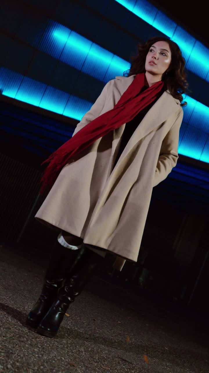 Inquadratura cinematografica a figura intera di una donna in piedi di notte in un ambiente urbano, mentre il vento le scompiglia i capelli e la sciarpa rossa sotto l'illuminazione al neon blu. Concetto per la moda street invernale, la fiducia e la vita notturna.