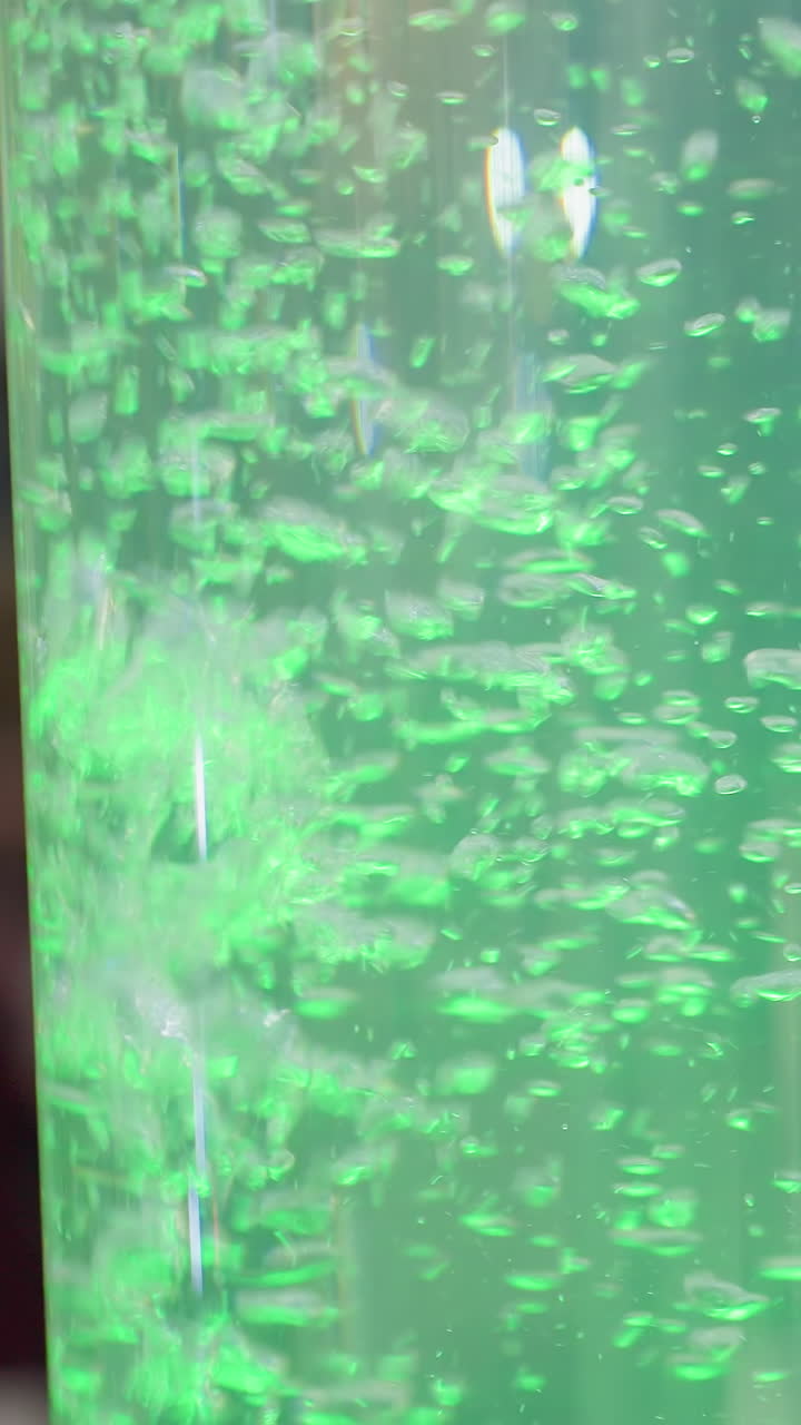 Close-up of large cylindrical water feature with green glowing water and floating bubbles, soft blur background and vibrant light effects create a tranquil, aesthetic atmosphere