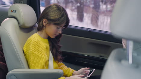 camera focuses on girl sitting on the back seat using a tablet while talking to their parents