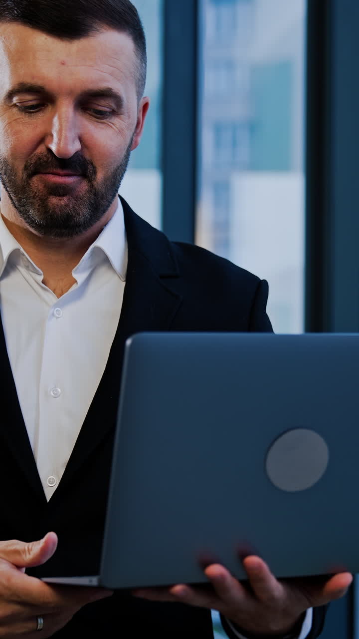 Smiling good-looking bearded businessman in suit stands near the window holding laptop. Man talks showing something with his hands. Vertical video