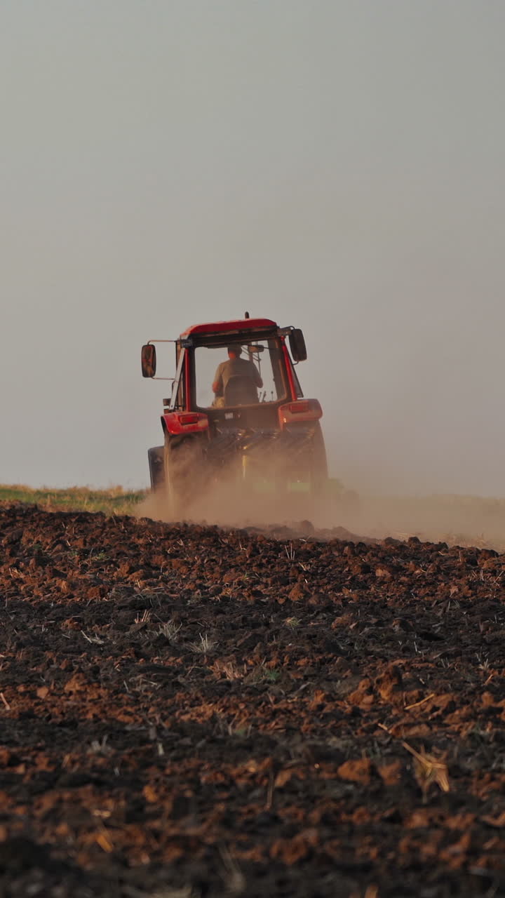 Brown field background and a tractor plowing the soil. Tractor cultivating the field, preparing land for sowing in the farmland. Vertical video
