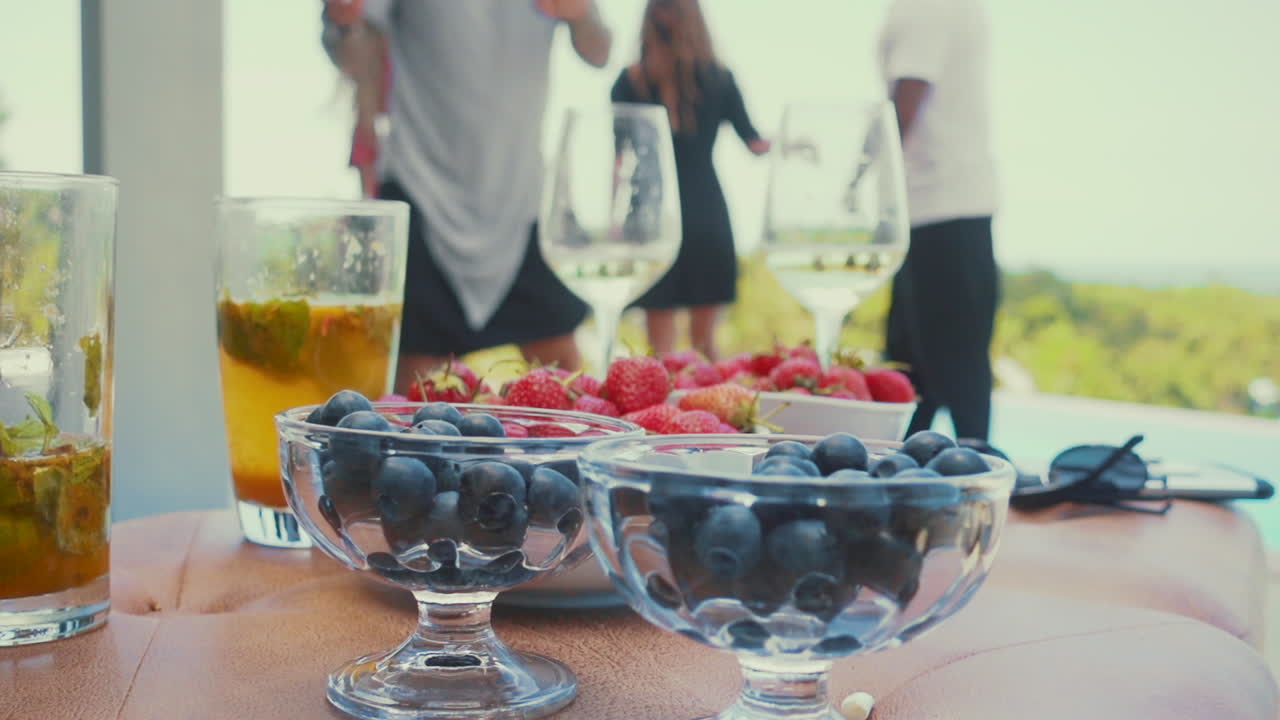 Sløret baggrund med mænd og kvinder der danser og hygger sig i en poolvilla, med drinks og cocktails med jordbær og blåbær i forgrunden
