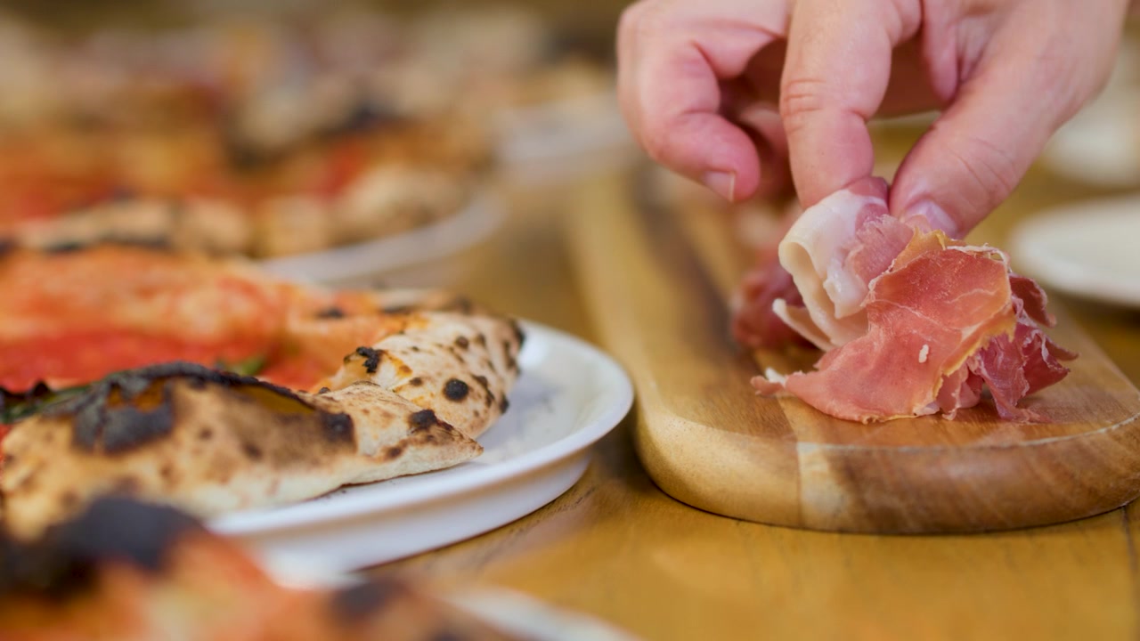 Primo piano di una mano che solleva salumi da un tagliere di legno accanto a pizze italiane fresche