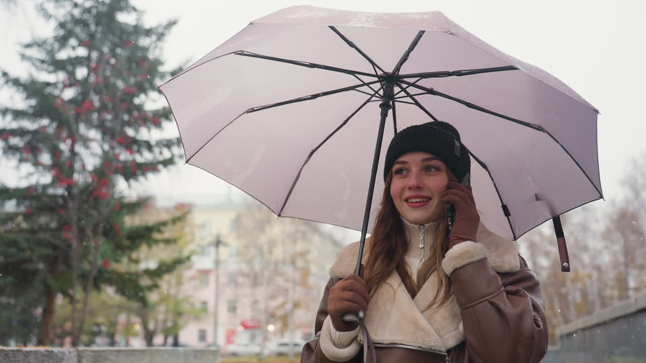 Dreamy girl smiling on phone call holding umbrella, wearing knit cap, brown shearling jacket, walking in light snowfall with soft background of autumn trees and urban setting