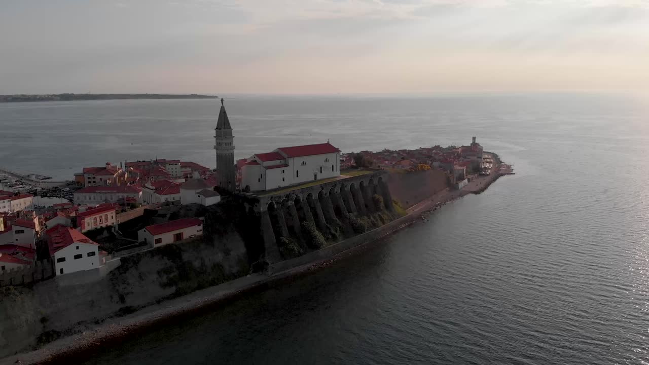 Ancient catholic church on top of the cliff with sunset in the background. Piran, Slovenia. Ancient tower with big clock on the Peninsula. Seagulls fly around the cathedral.