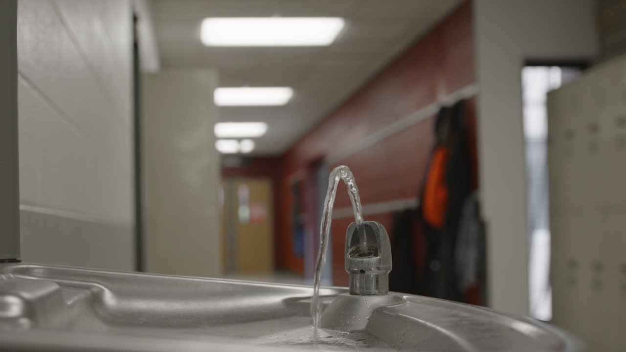 A quiet high school or middle school hallway featuring a running water fountain in focus with coats hanging out of focus in the background—capturing the stillness and realism of daily school life.