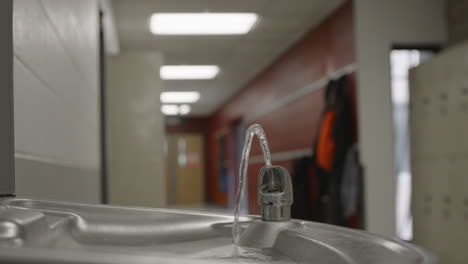 a quiet high school or middle school hallway featuring a running water fountain in focus with coats hanging out of focus in the background—capturing the stillness and realism of daily school life.