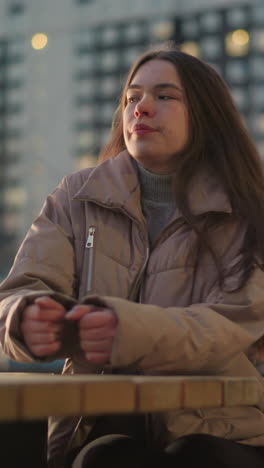 a young woman dressed in a peach jacket and black trousers is seen sitting on a bench in an urban park. she places her hand on the table and gazes off into the distance with a thoughtful expression
