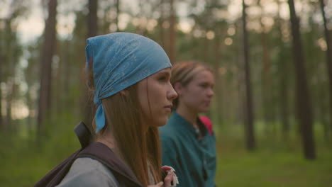 close-up of ladies walking along forest trail surrounded by tall trees, one swings her black bag over her shoulder while gazing warmly, while the other walks beside her softly blur in the background