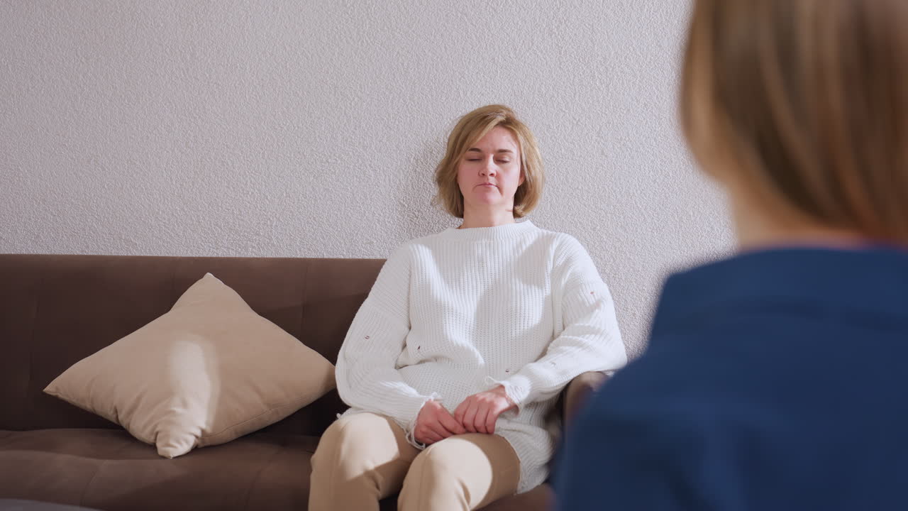 Guest seated on brown sofa opposite therapist listening intently and nodding in response during therapy session pillow at side, neutral wall background and soft warm lighting in cozy counseling room