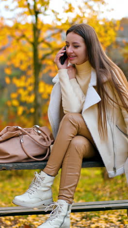 calm relaxed lady in white jacket and beige leggings speaks on her phone. resting woman spending time in the autumn park sitting on the wooden table. vertical video