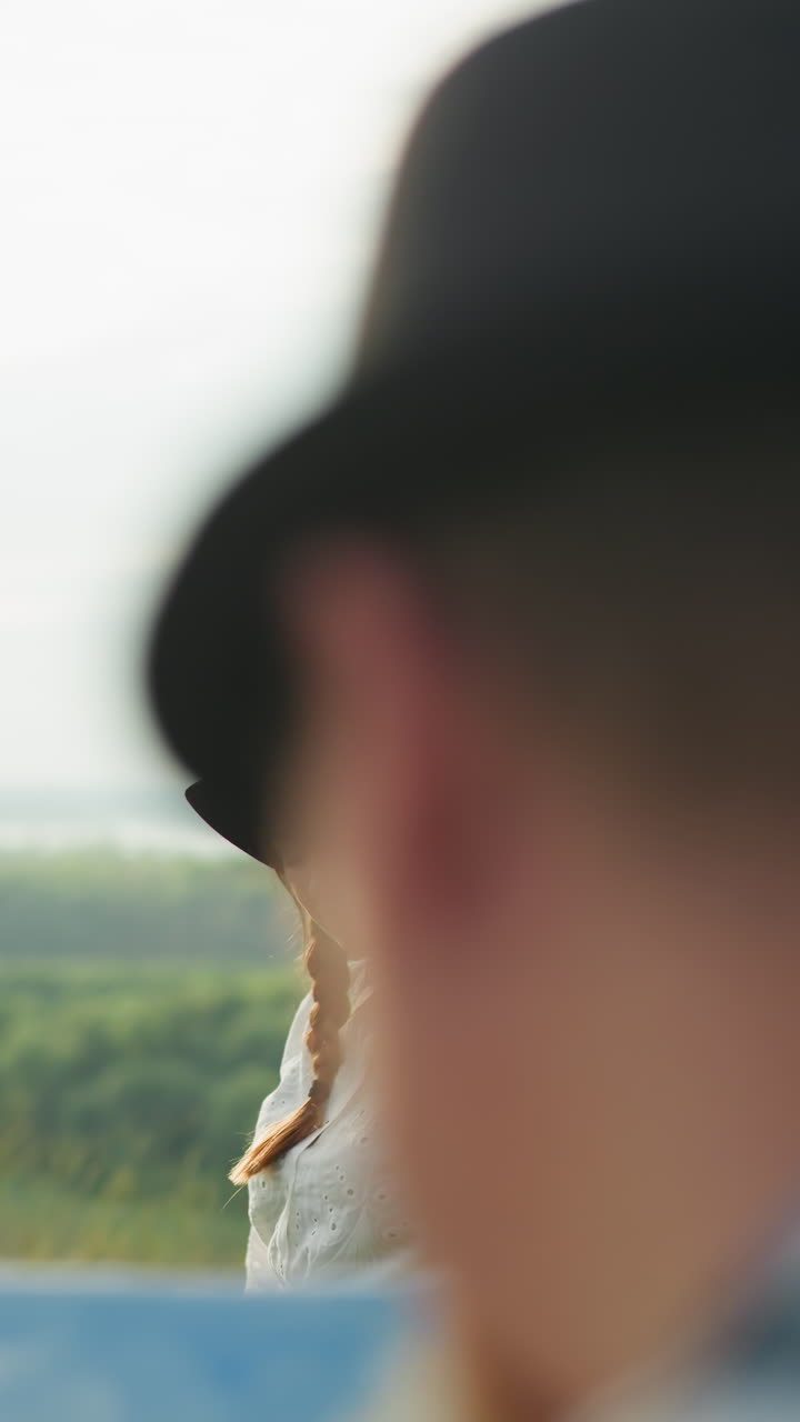 색 드레스와 모자를 입은 여자가 조용한 호수를 내려다보는 잔디에 우아하게 포즈를 취하고, 앞면에는 모자를 입고 있는 남자의 흐릿한 클로즈업이 있습니다.