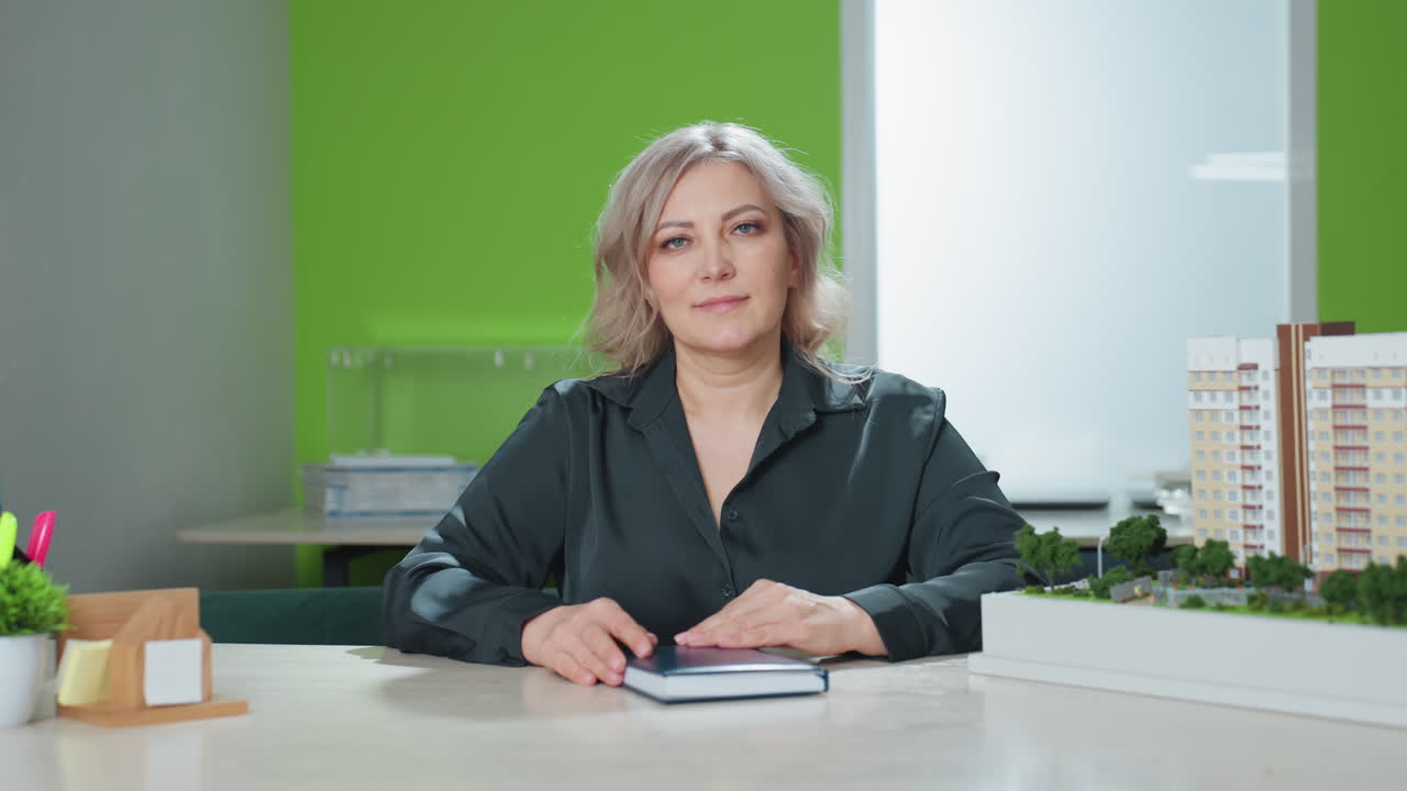 woman in professional attire seated at desk looking confidently into camera with hands resting on closed notebook, positioned beside architectural model in bright modern office