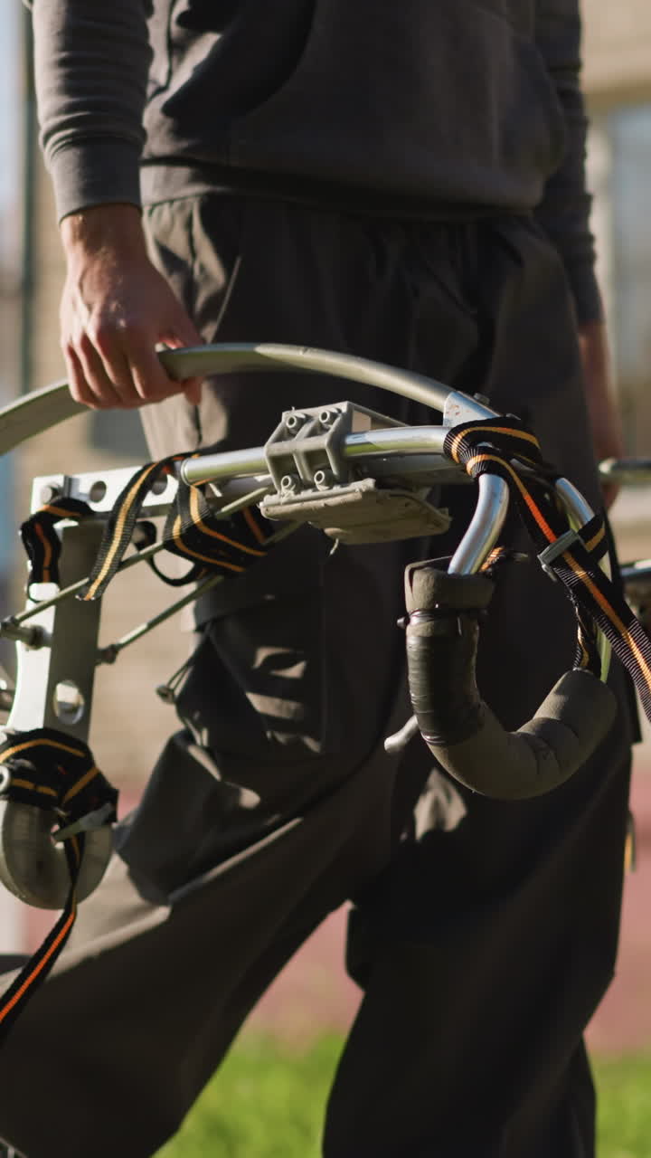 persona sosteniendo pilotes metálicos con mangos y correas oscuras en el exterior cubierto de hierba, con sudadera gris oscuro y pantalones sueltos, sólo las manos y la parte inferior del cuerpo visibles, el fondo borroso con hierba y edificio