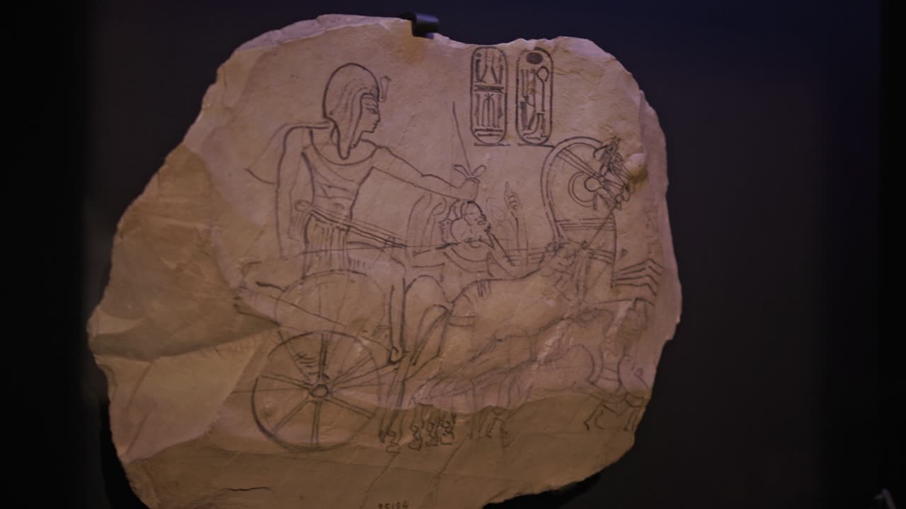 Primo piano di un antico disegno egizio raffigurante un guerriero su un carro trainato da cavalli, abbozzato su un frammento di papiro o calcare con geroglifici visibili.