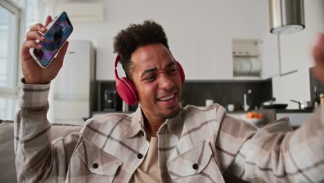 Happy-young-man-with-Black-skin-color-brunette-in-red-headphones-and-a-cream-plaid-shirt-listens-to-cheerful-music-along-with-his-phone-in-his-hands-while-sitting-on-a-brown-sofa-and-dancing-in-a-modern-apartment