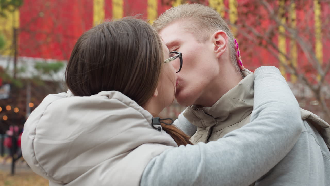 Newly married couple sharing affectionate kiss outdoors while embracing closely in casual jackets, man gazes at wife lovingly with people walking in background