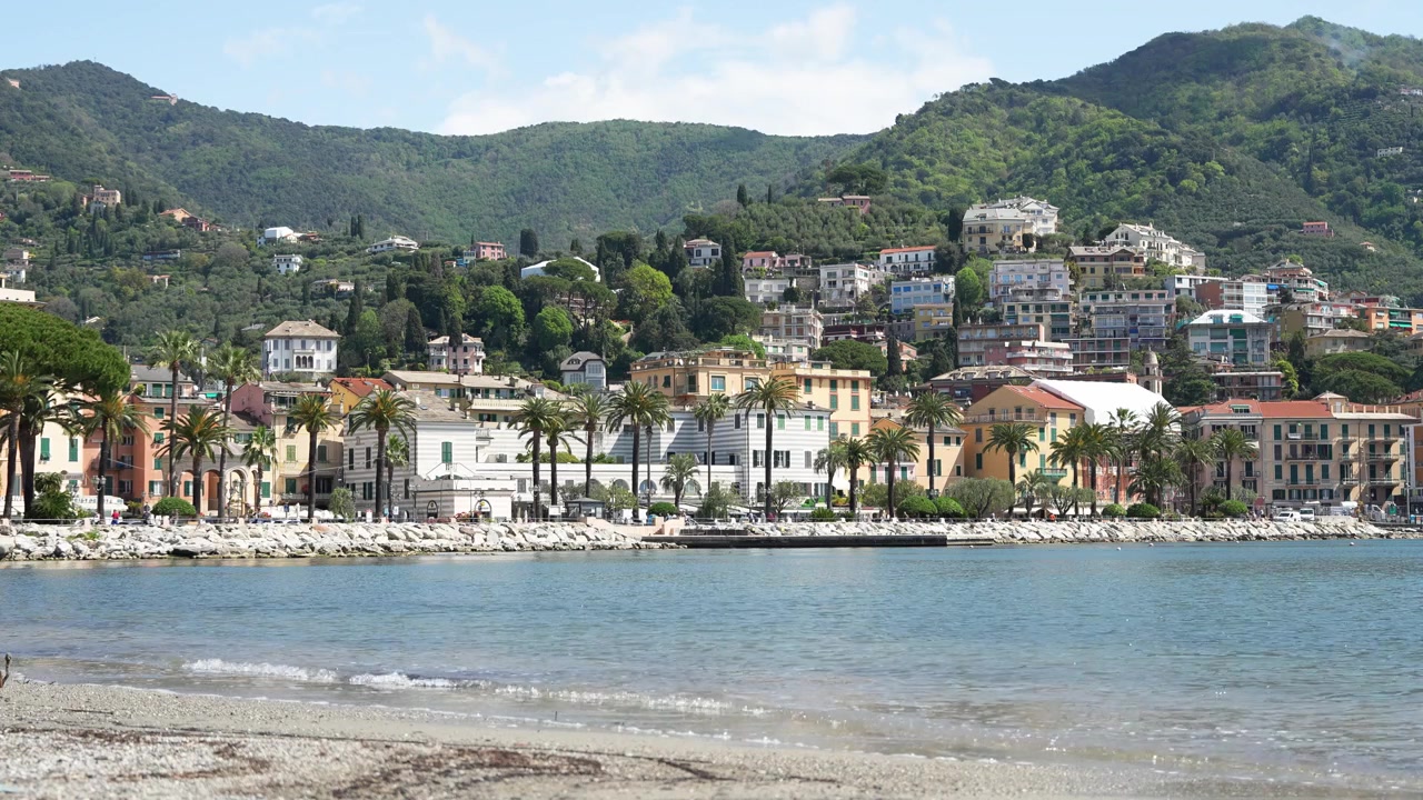 Inquadratura grandangolare di Rapallo, Italia, che mostra vivaci edifici sul mare, palme, acqua blu tranquilla e colline rigogliose sotto la luce del sole. Ideale per filmati di repertorio di viaggi o turismo.
