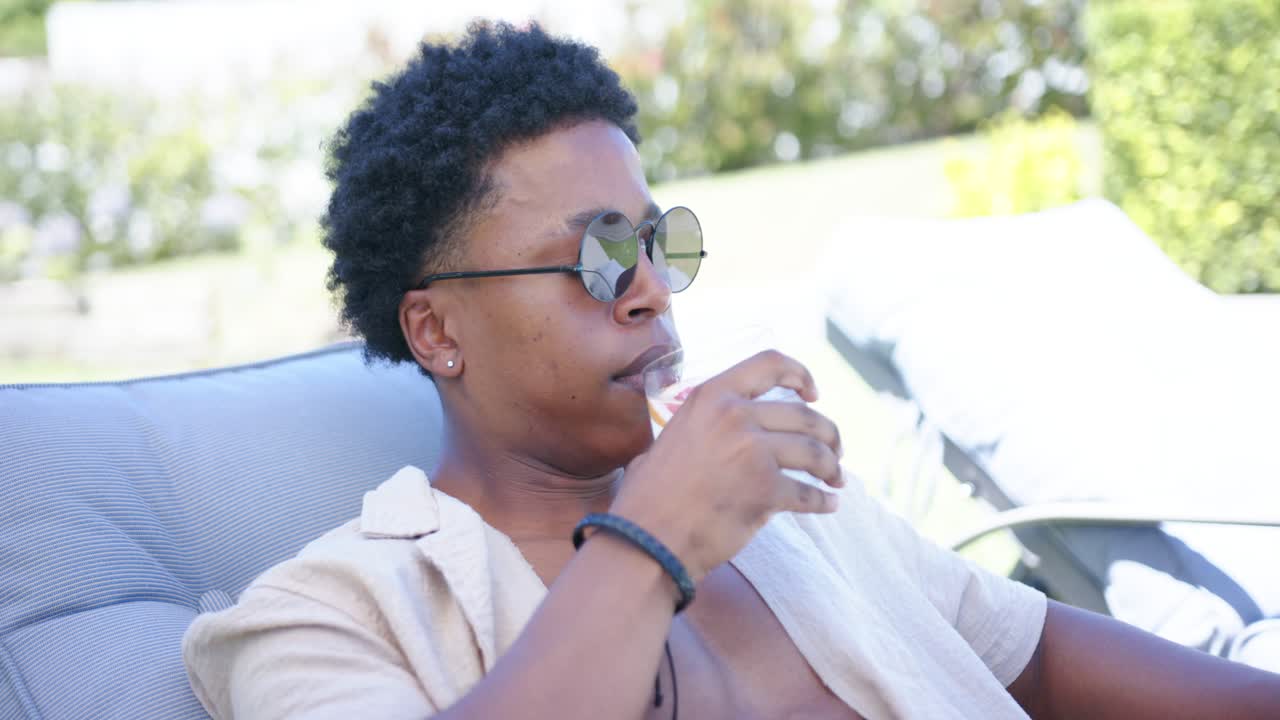 Afroamerikansk mand læner sig tilbage i en stol i lyserøde badebukser, holder et glas og nipper for at køle ned ved poolen