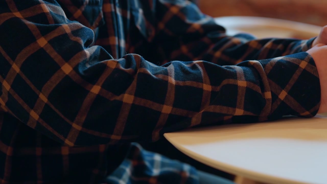 Primo piano di una persona con camicia a quadri con un tavolo di legno e un piano bianco che si concentra sulle mani che sistemano oggetti. L'illuminazione soffusa evidenzia i dettagli della texture come le venature del legno o il tessuto.