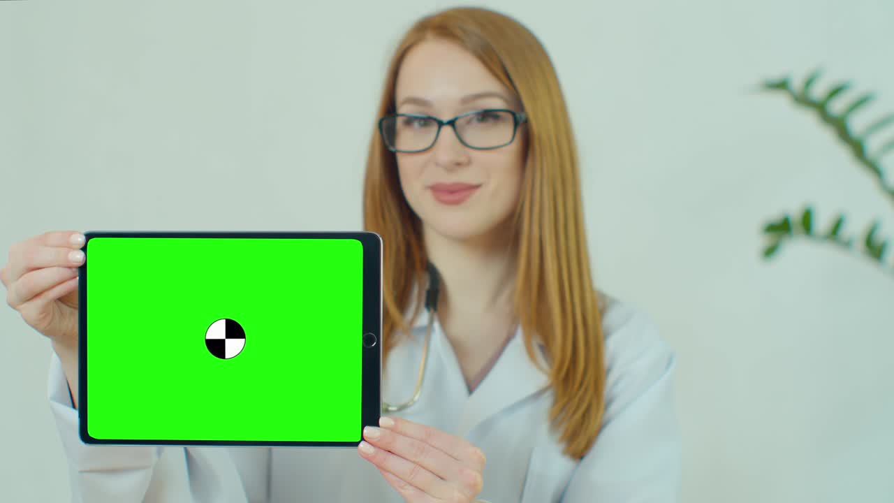 Female Doctor in glasses demonstrating digital tablet with chroma key in hospital, Portrait of woman in white gown with stethoscope in medical office showing digital tablet with green screen.
