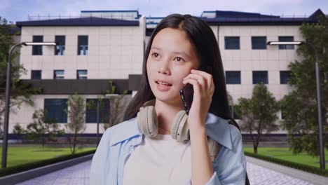 close up of asian teen girl student with a backpack talking on smartphone while standing in front of a school building