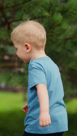 toddler child in blue t-shirt stands surprised when pine cone strikes boy arm in eco hotel yard in coniferous forest close side view slow motion