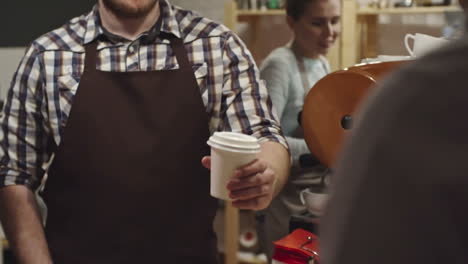 smiling barista in apron giving customer coffee in paper cup and thanking him for order at coffee shop counter