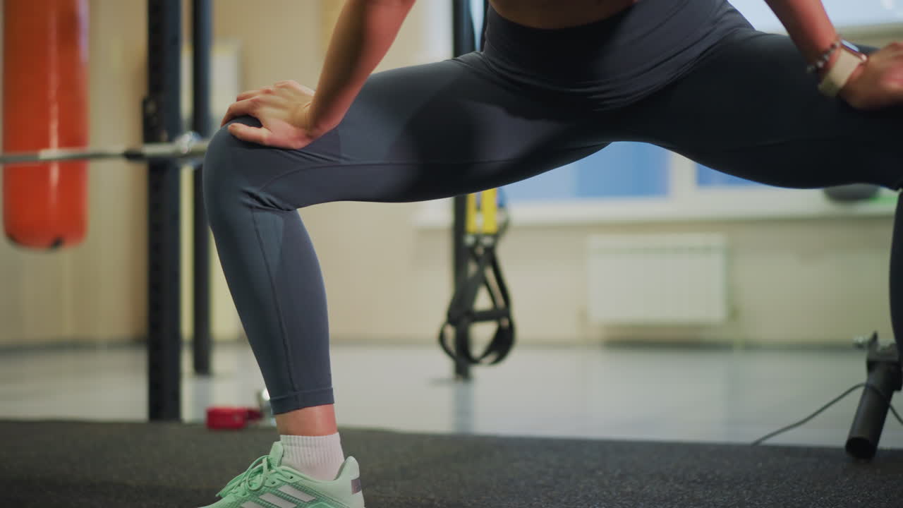 Primo piano in palestra di gambe con scarpe da ginnastica che eseguono affondi laterali profondi, atleta donna in leggings grigi, postura controllata, respirazione concentrata, squat rack e fasce di resistenza sullo sfondo, pavimento con tappeto da palestra
