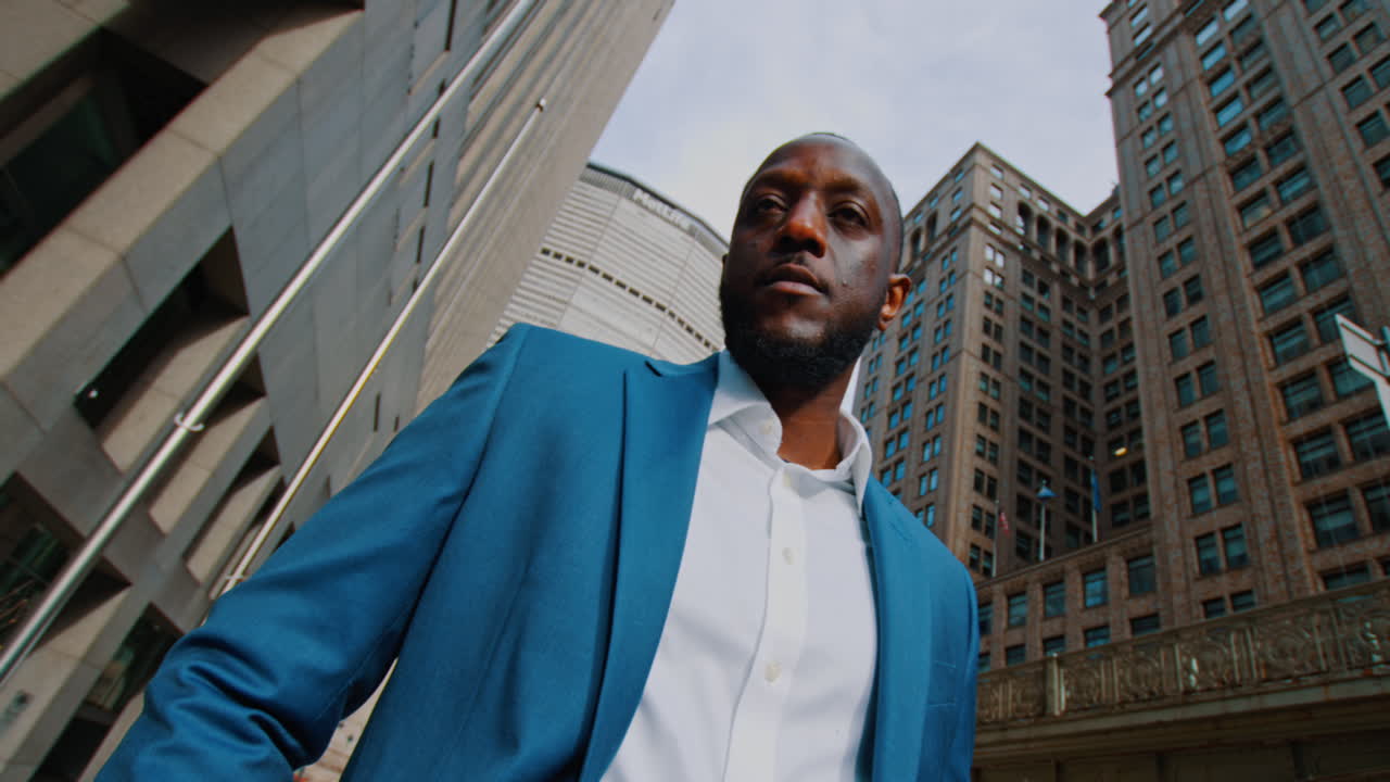 New York, USA, 8 April 2026: Low angle shot of businessman among New York skyscrapers. Worms eye view of African American man in blue suit looking down at camera against city buildings