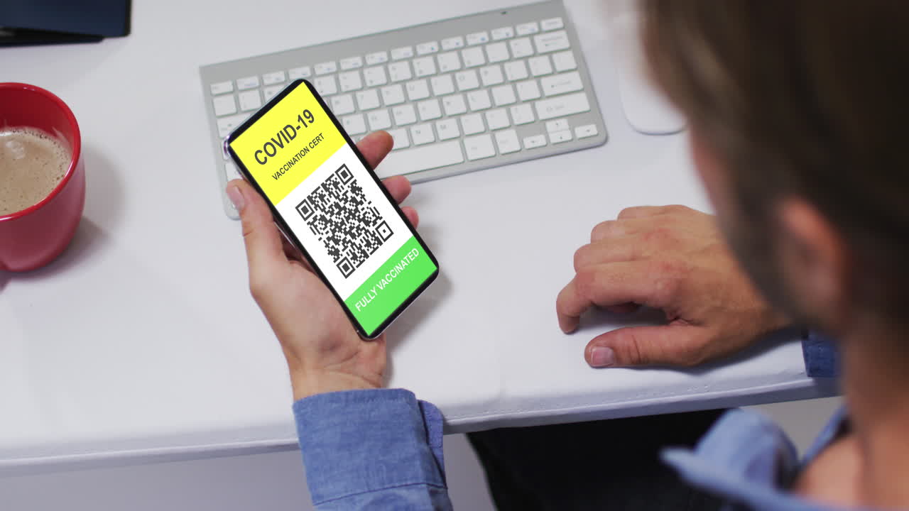 Man at desk holding smartphone with covid vaccination certificate and qr code on screen