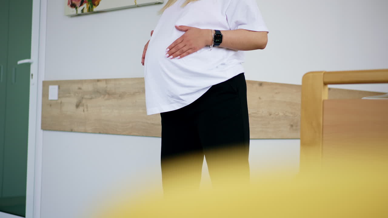 Unknown pregnant lady wearing white t-shirt and black pants. Woman touches her belly and walks by the ward.