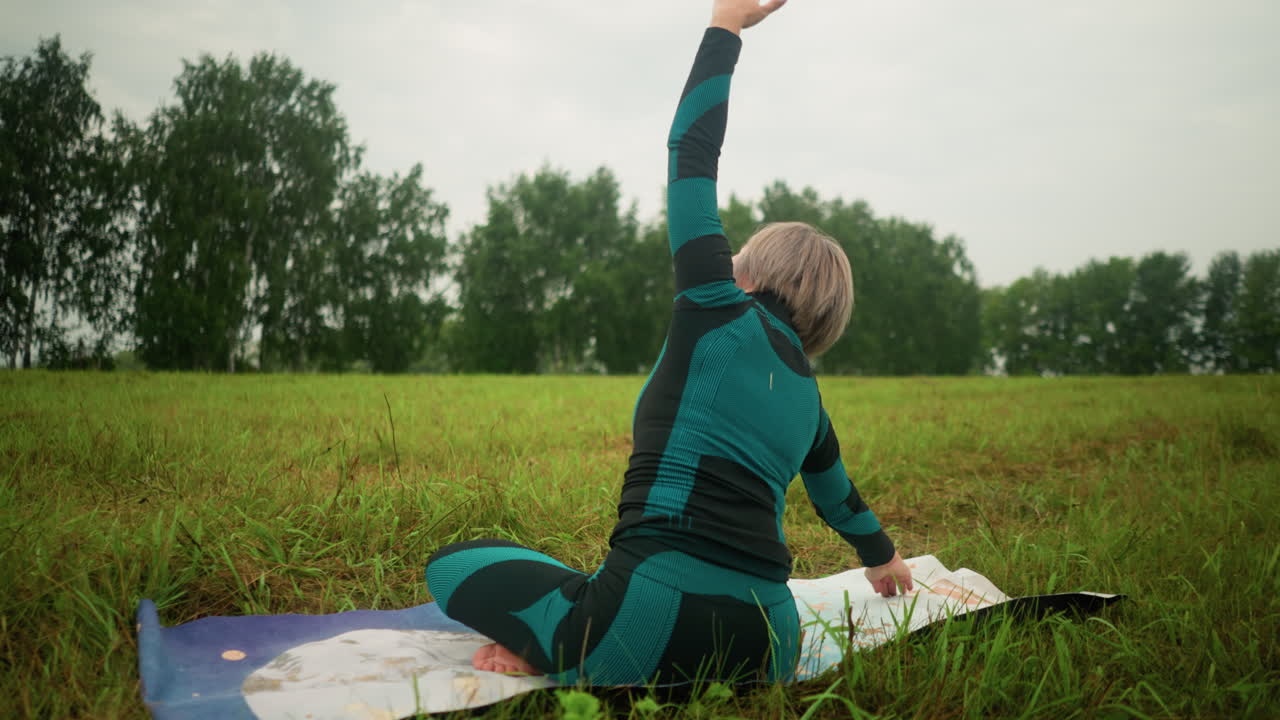 vista trasera de una mujer acostada en una alfombra de yoga, practicando la postura de flexión lateral con los brazos extendidos, y sentada en una postura lograda bajo un cielo nublado, en un vasto campo de hierba con árboles en la distancia