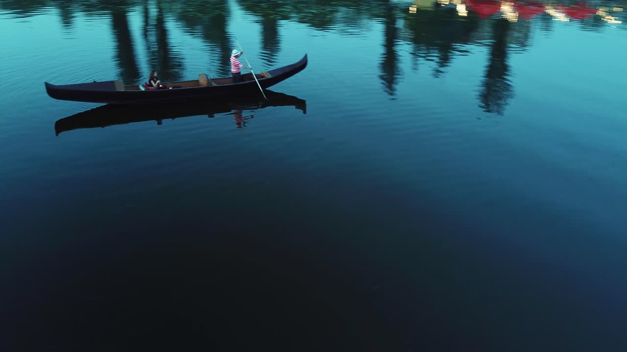 Woman and a gondolier floating in a boat on the evening river. Man in white hat and striped shirt carring elegant woman with lantern in a boat. Quick aerial view around.