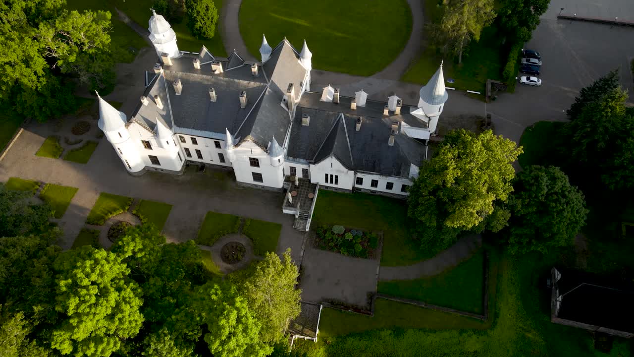 Gorgeous aerial drone footage flying closer at high altitude to a white colored majestic and historic Alatskivi castle during sunset and tilts the footage down to go to a top down shot during summer