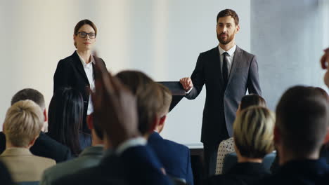 relatrice caucasica che tiene un discorso a una conferenza e risponde alle domande dell'uditorio, il suo collega maschio in piedi accanto a lui sul podio