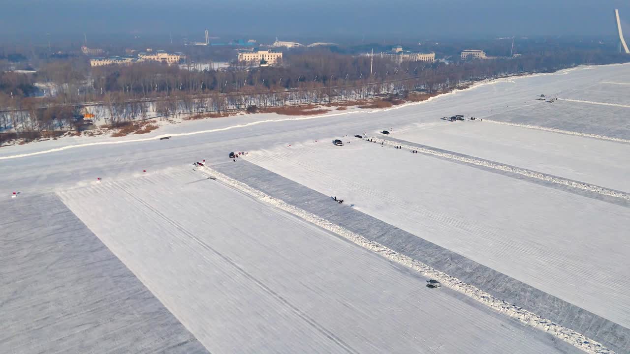 Aerial view of ice-cutting machines at work on the frozen Songhua River in Harbin, creating parallel cuts in the ice. The vast, snow-covered surface is prepared for the ice blocks. China