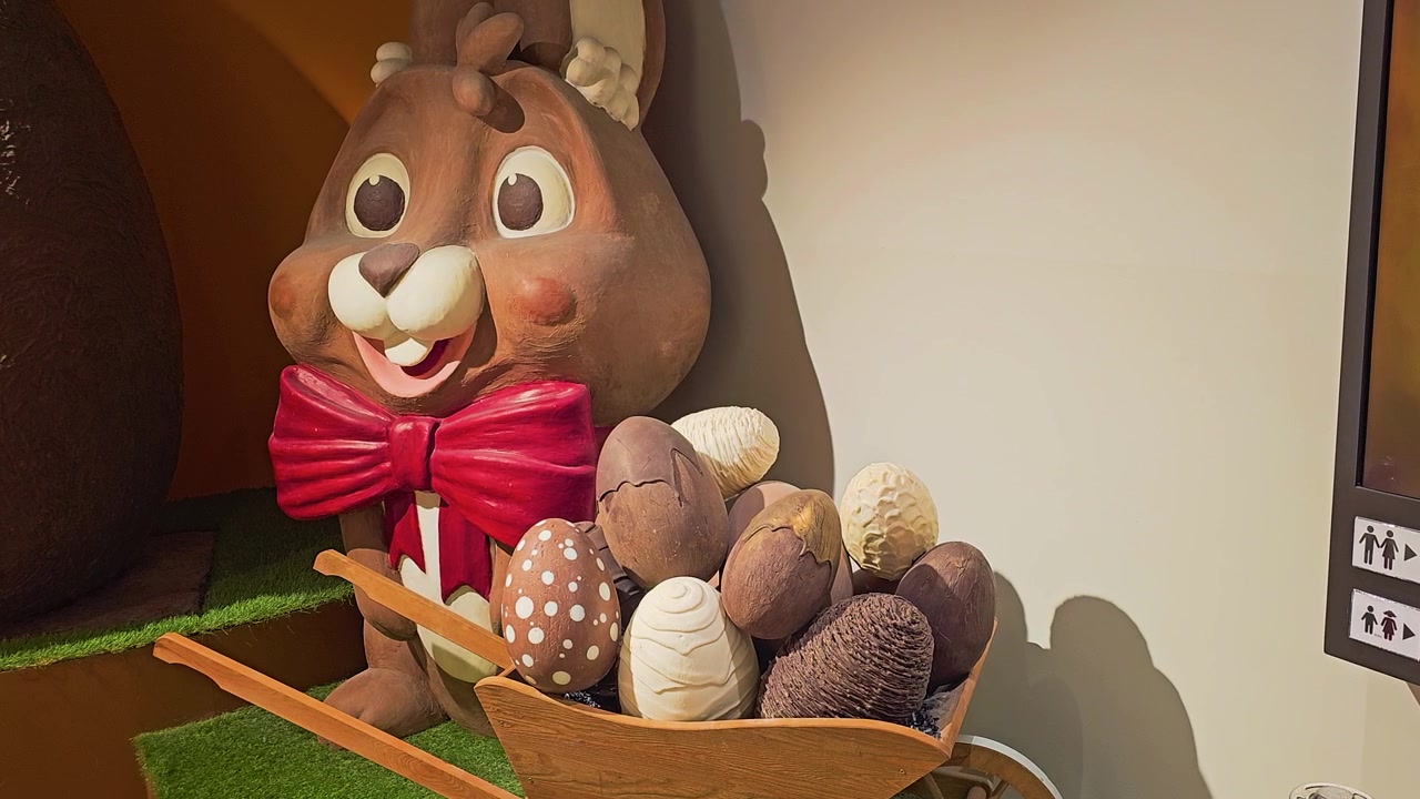 Decorative Easter bunny display with a large smiling rabbit, red bow tie, wooden cart full of colorful eggs, warm indoor lighting, and festive chocolate shop or holiday store atmosphere.