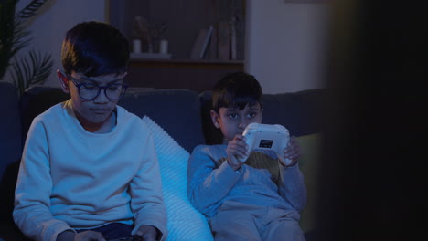 Two-Young-Boys-Sitting-On-Sofa-At-Home-Playing-With-Computer-Games-Console-On-TV-Holding-Controllers-Late-At-Night-1