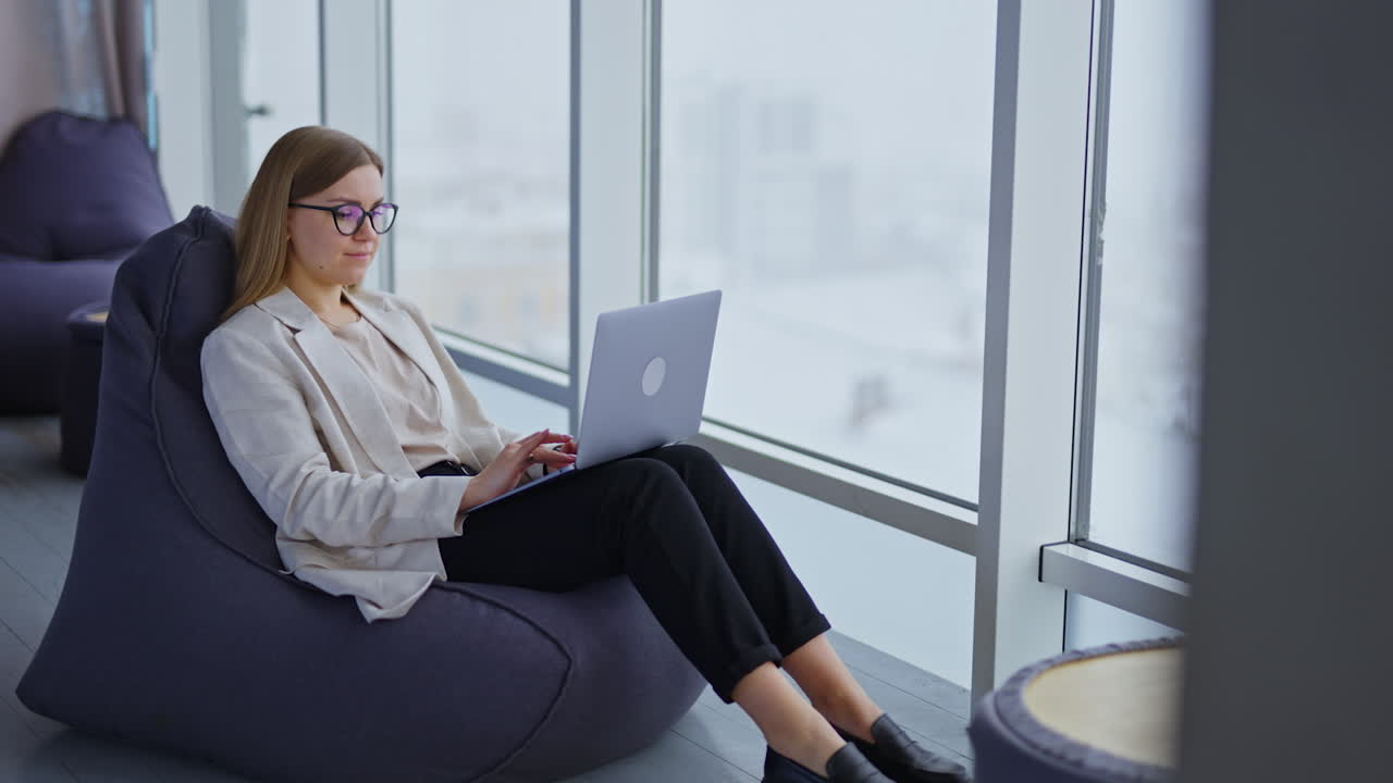 Freelance female entrepreneur working at laptop. Lady sits in comfortable chair near the big panoramic window.