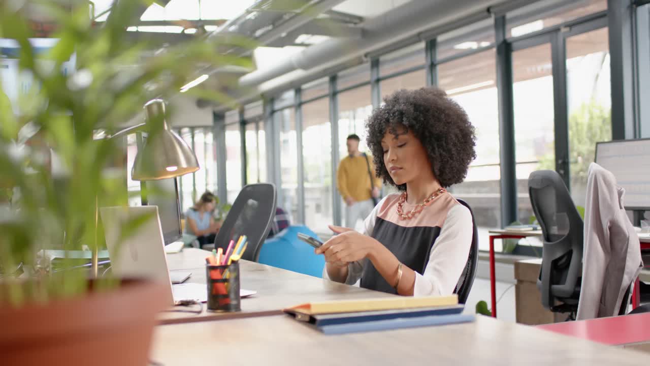 mulher afro-americana usando um smartphone sentada em sua mesa no escritório